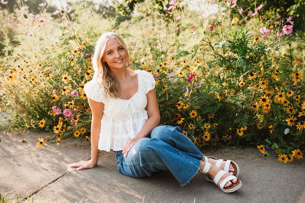 Young blonde woman sitting outdoors in jeans and white top, surrounded by colorful wildflowers in golden sunlight.