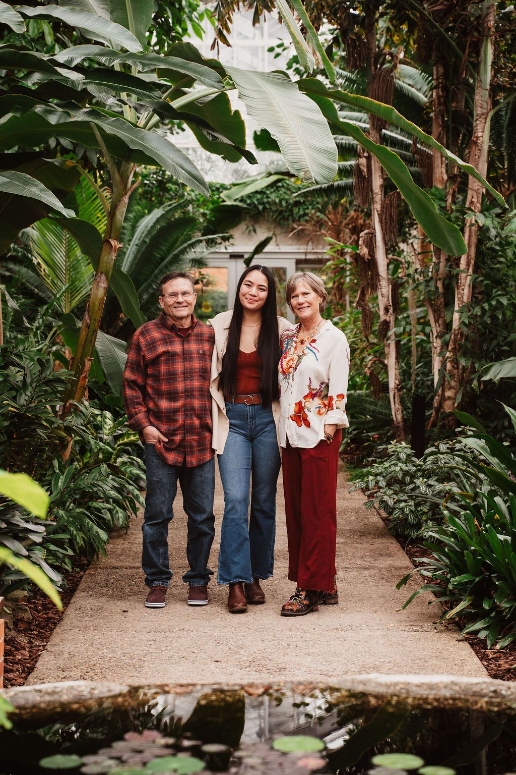 A young woman stands with her parents on a garden path surrounded by lush tropical plants.
