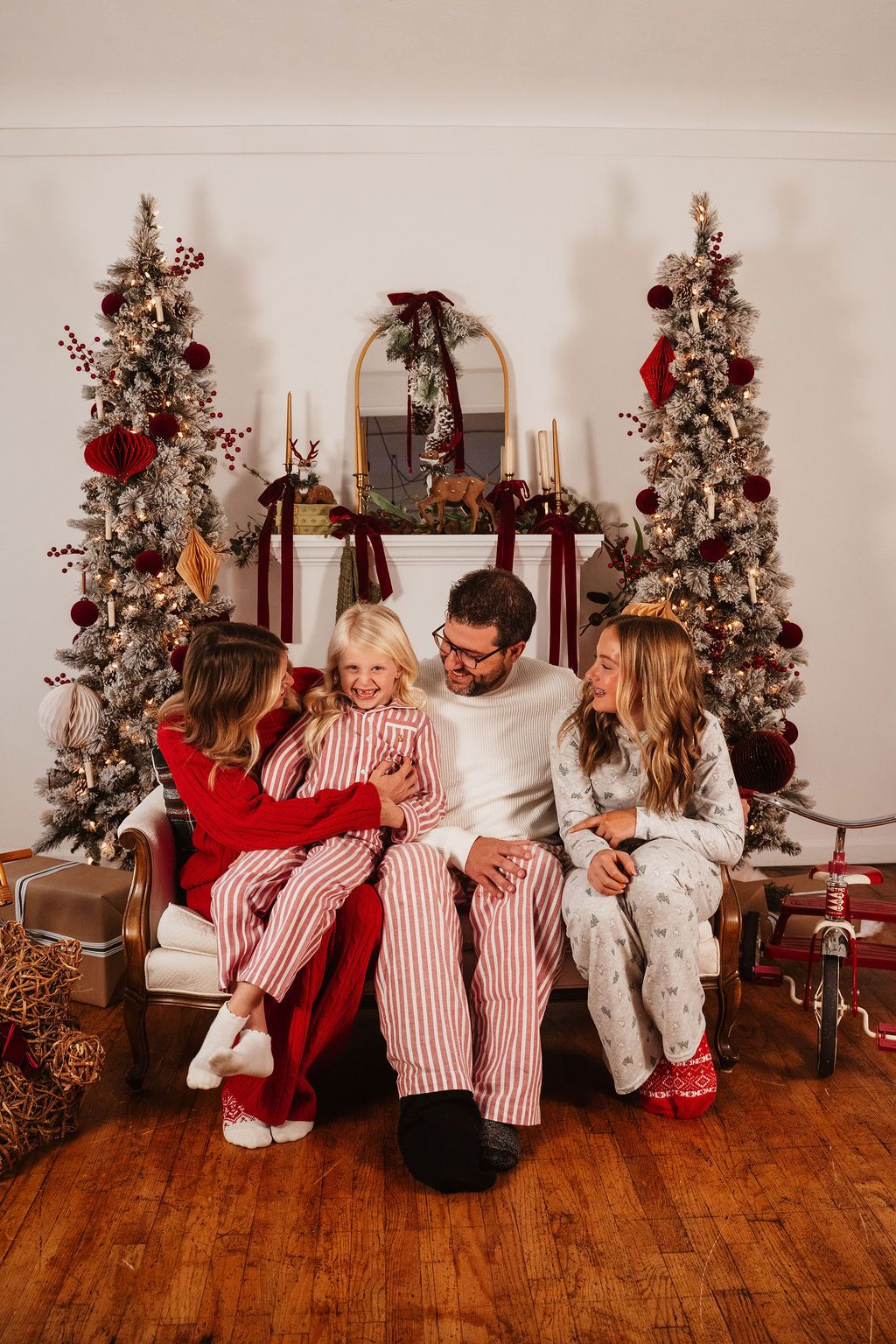 Family of four in matching Christmas pajamas sitting together by decorated holiday trees and fireplace.