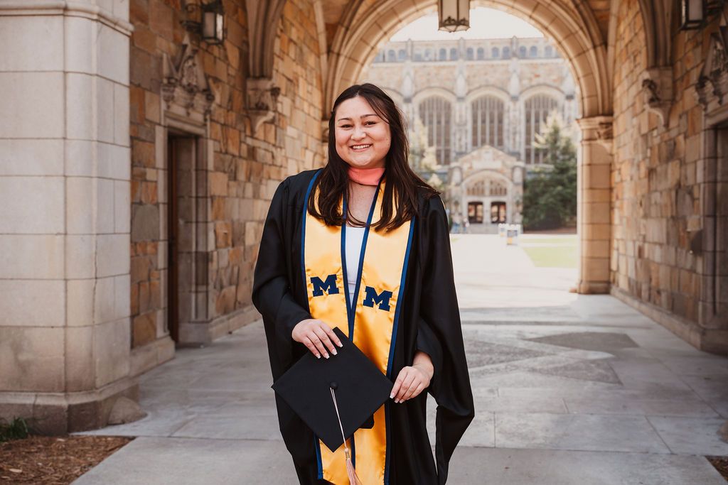 Graduate in black gown with two maize Michigan stoles walks under campus archway at U of M.