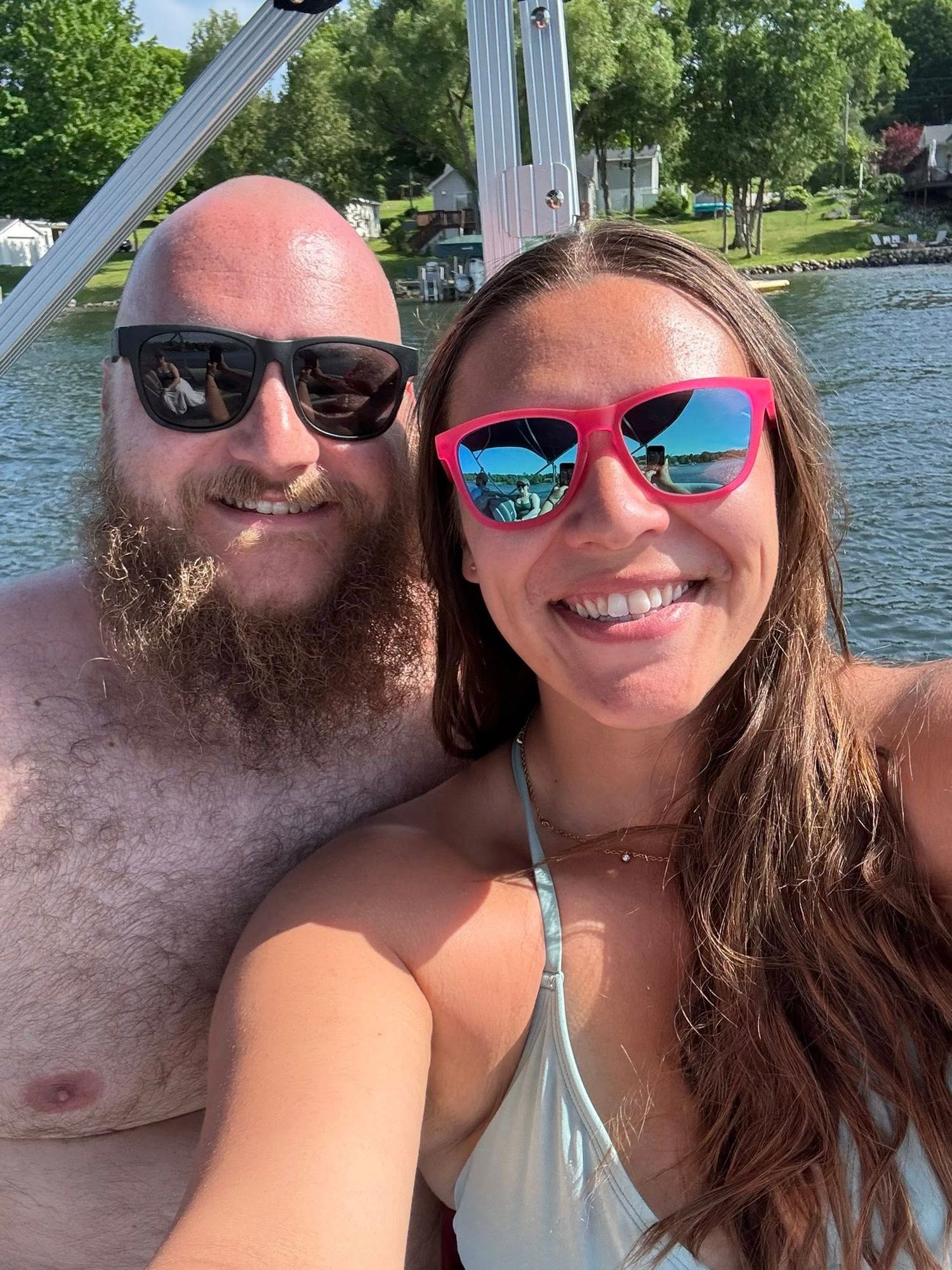 Couple taking a selfie on a boat with sunglasses, smiling with a lake and green trees in the background.