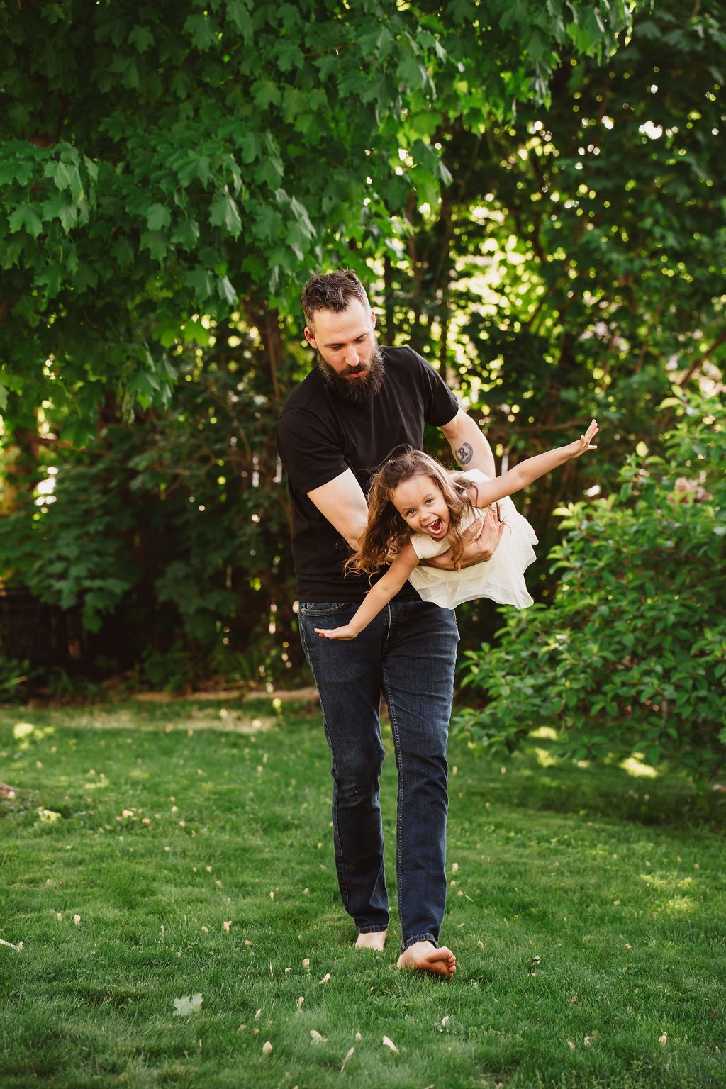 Father playfully spinning laughing daughter outdoors on lush green lawn surrounded by trees on sunny day.