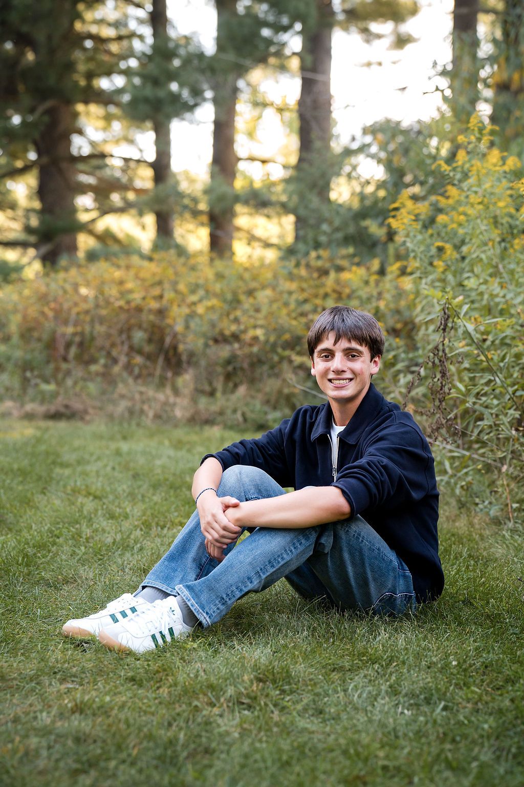 A person wearing a dark shirt and jeans lies casually on green grass in a wooded outdoor setting.