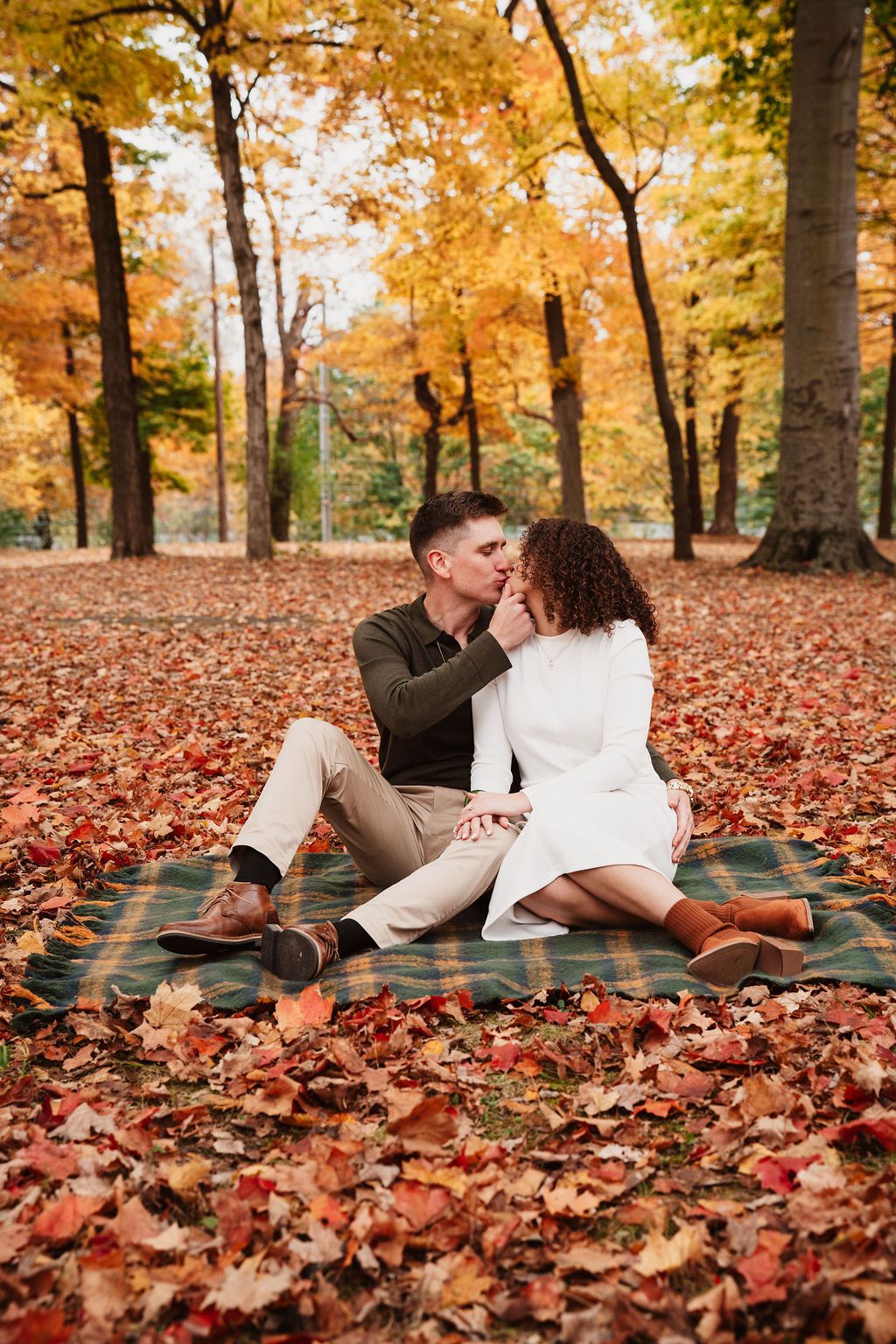 A romantic couple shares a kiss while sitting on a plaid blanket surrounded by vibrant autumn leaves in a park.