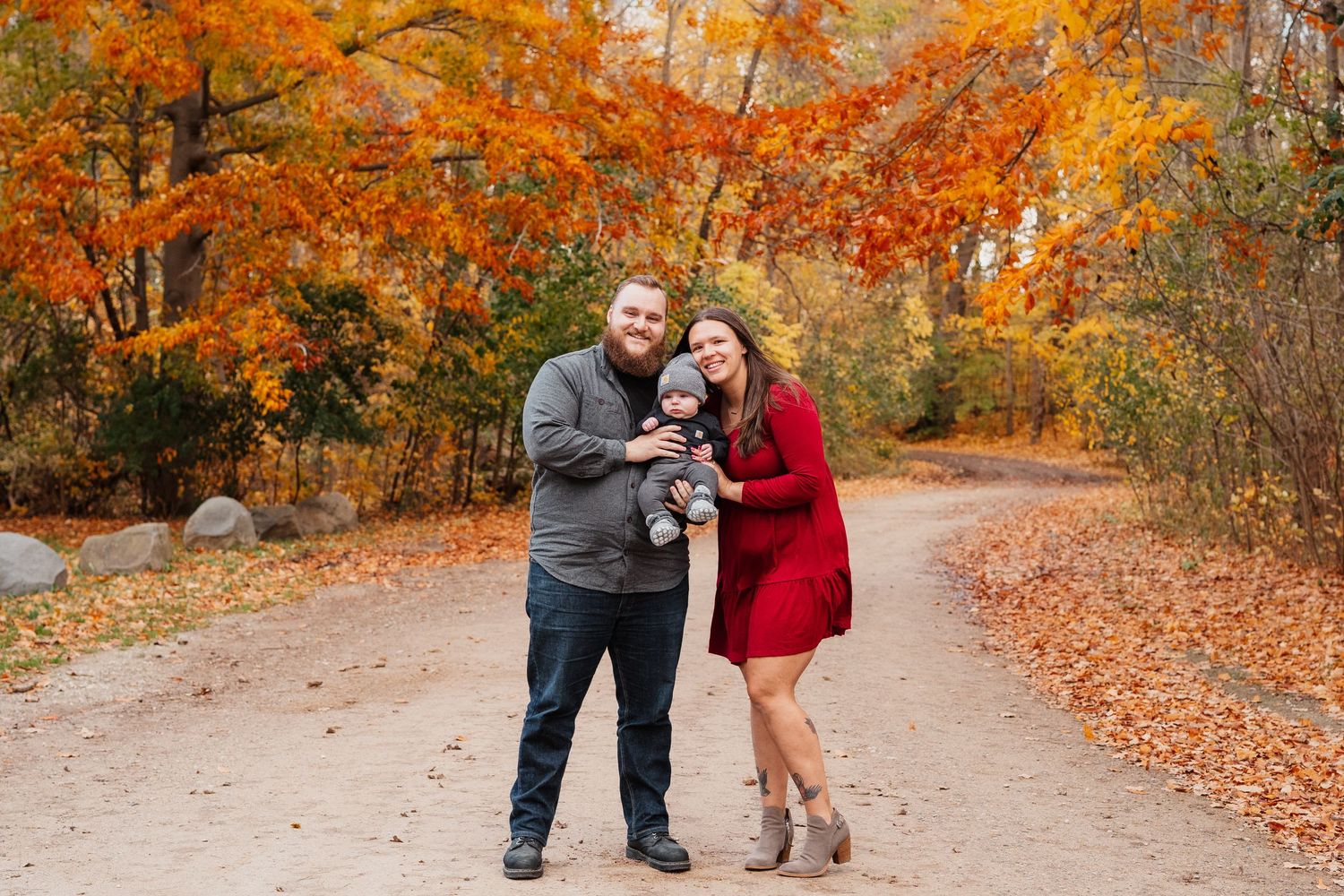 Family of three posing on autumn trail surrounded by vibrant orange and red foliage during fall season.