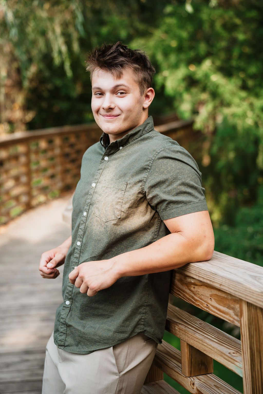 Young man in green shirt leaning on wooden bridge railing, smiling outdoors with lush greenery background.