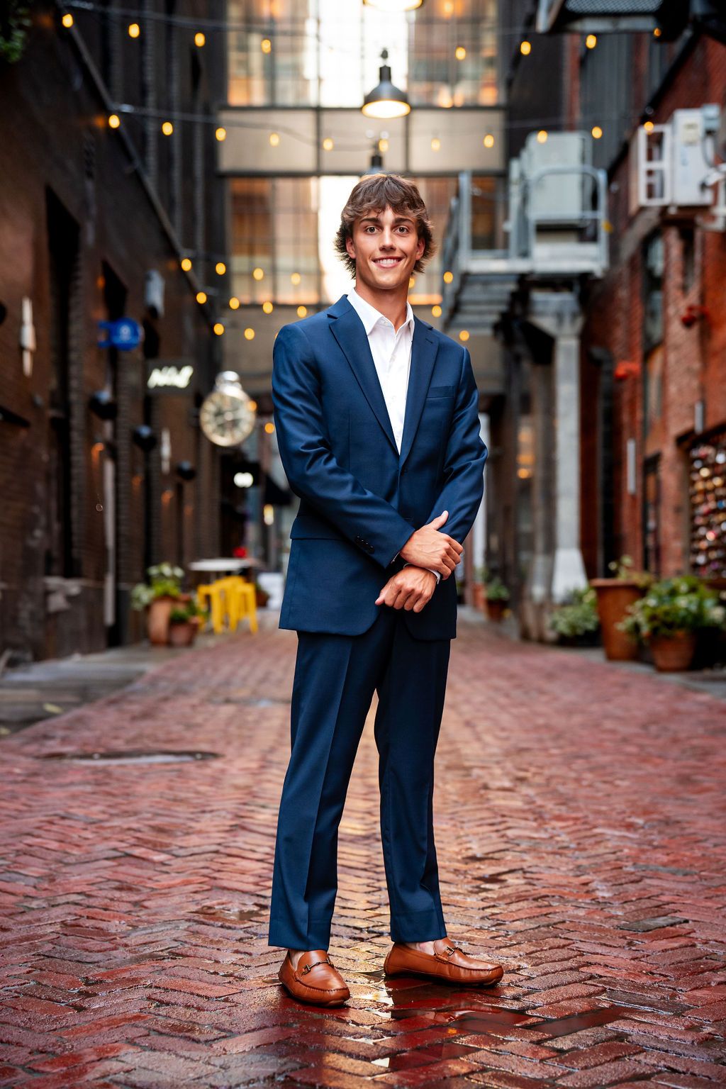 Young man in blue suit and loafers standing on wet brick alley with string lights.