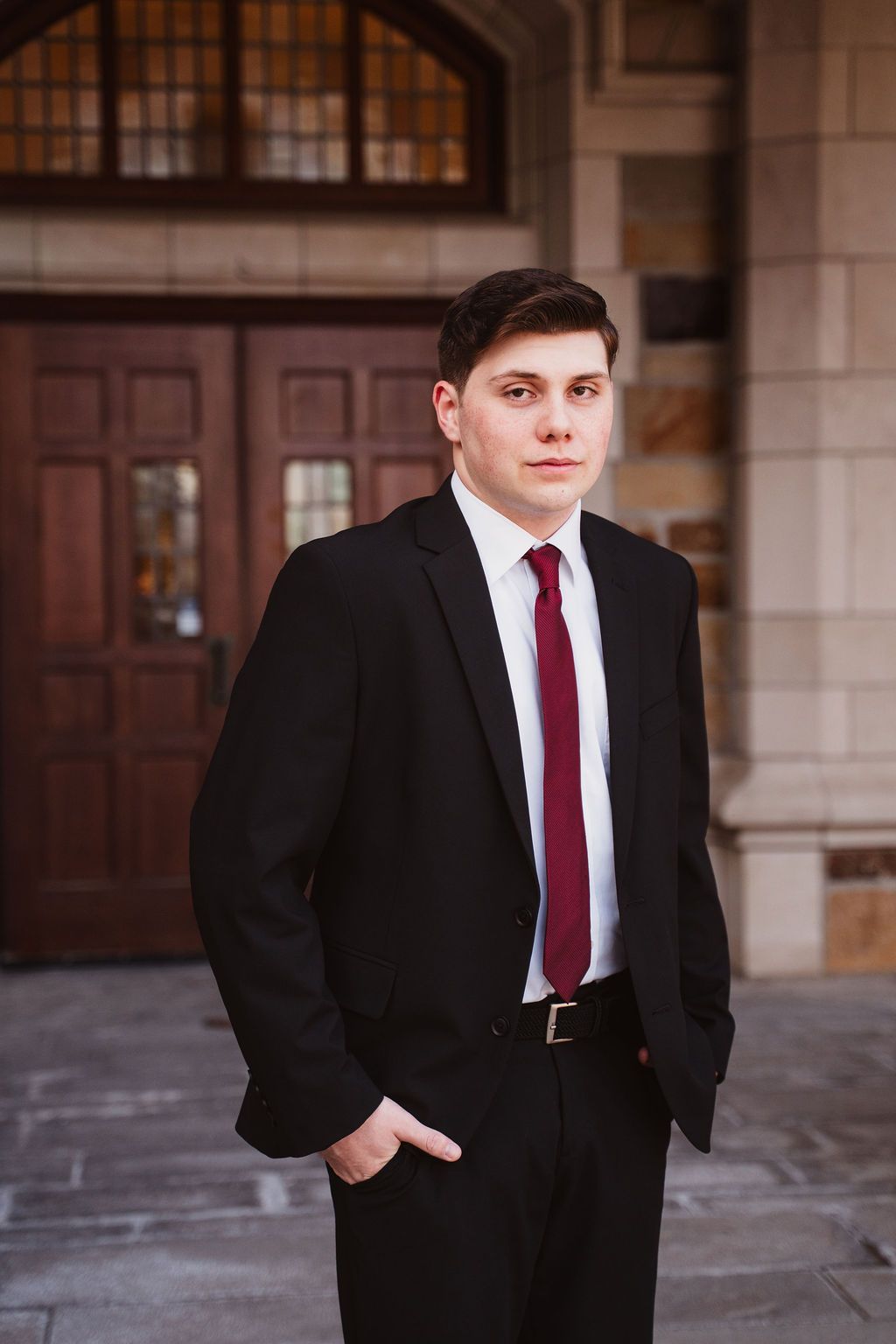 Young man in black suit and burgundy tie standing confidently outside a stone building with wooden doors.