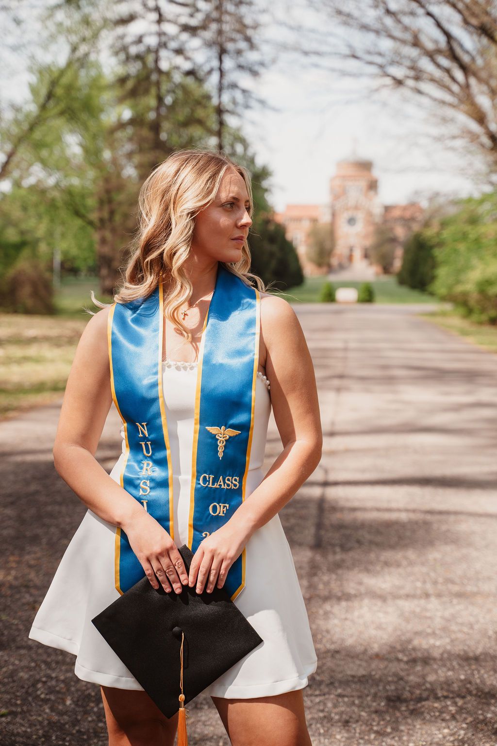 Nursing graduate poses outdoors in white dress with blue Class of 2024 sash, holding graduation cap.