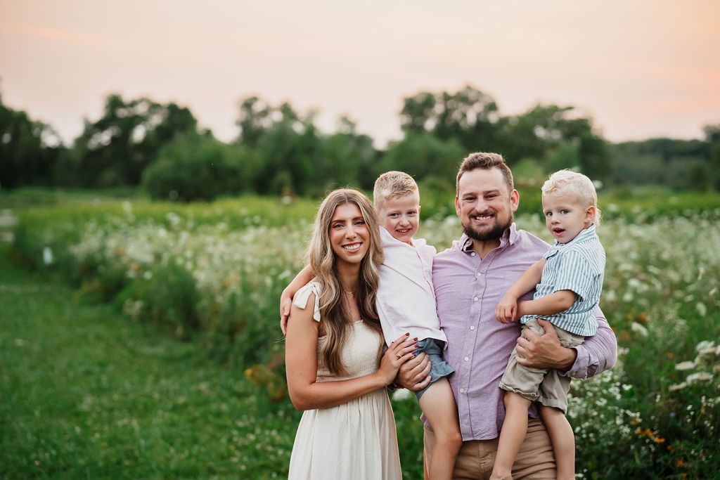 Happy family of four posing outdoors in a wildflower meadow at golden hour, parents holding two young children.