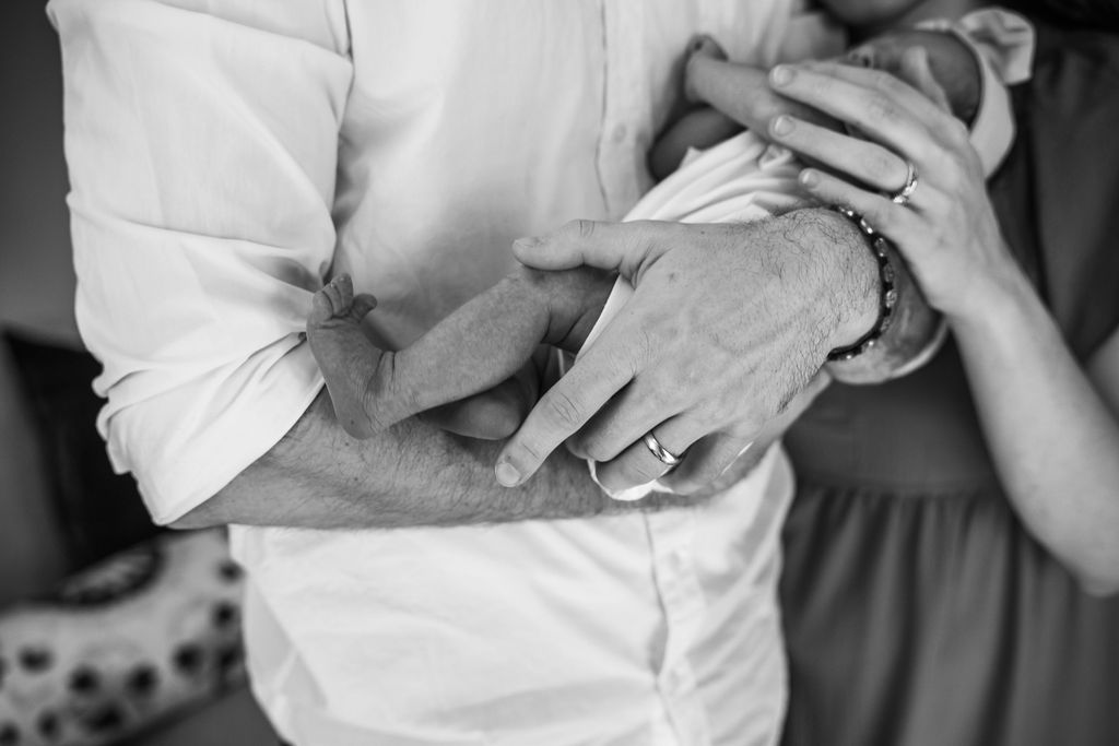 Black and white photo of parents cradling newborn baby, showing close-up of hands with wedding rings.