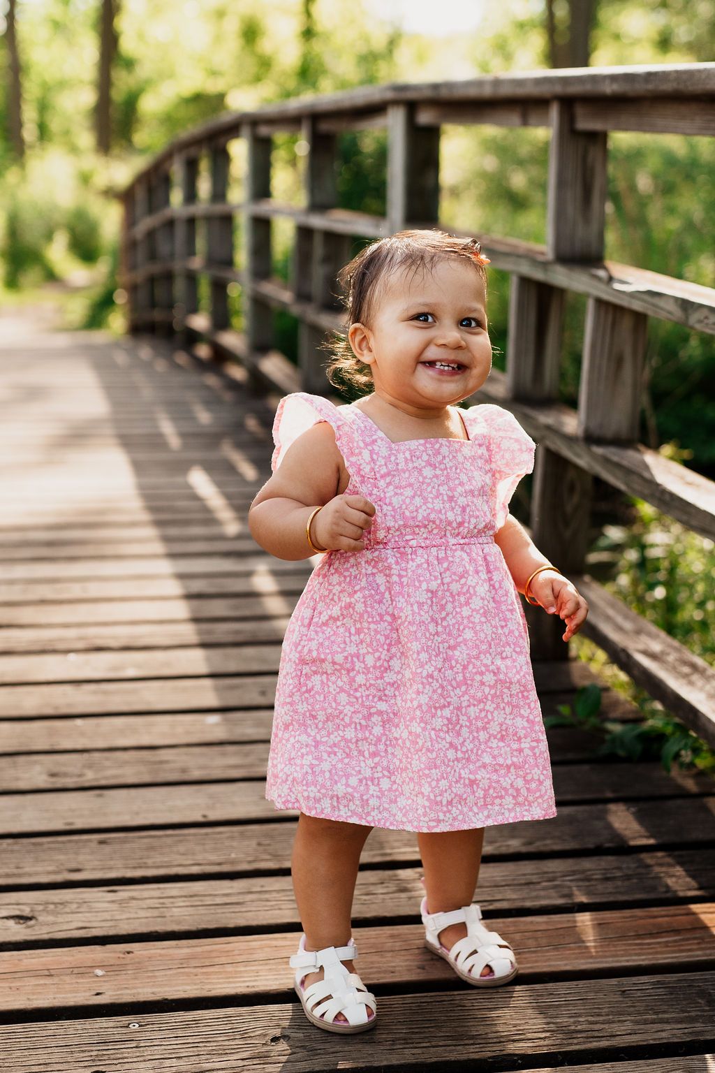 A small child wearing a pink floral dress and white sandals stands on a wooden boardwalk in a sunny outdoor setting.