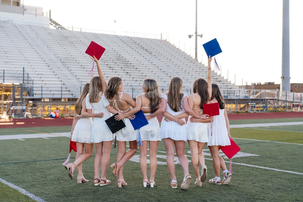 A group of cheerleaders in white uniforms throw graduation caps into the air on a football field.