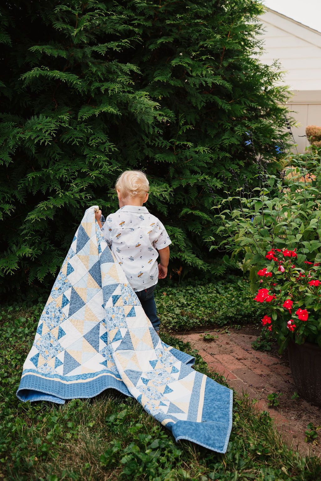 Blue and white geometric patterned quilt displayed in garden setting with evergreens.