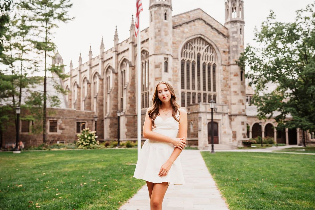 A person in a white dress poses in front of a Gothic-style university building on a sunny summer day.