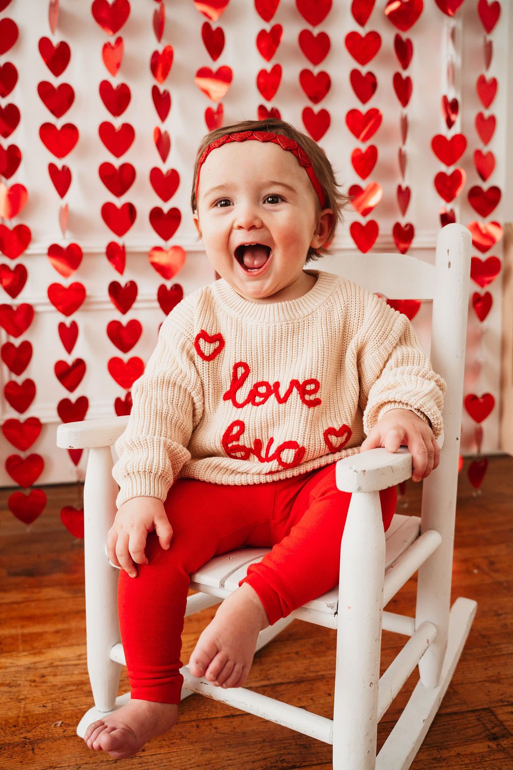 Happy baby in a love sweater and red leggings sits in a white rocking chair with Valentine heart backdrop.