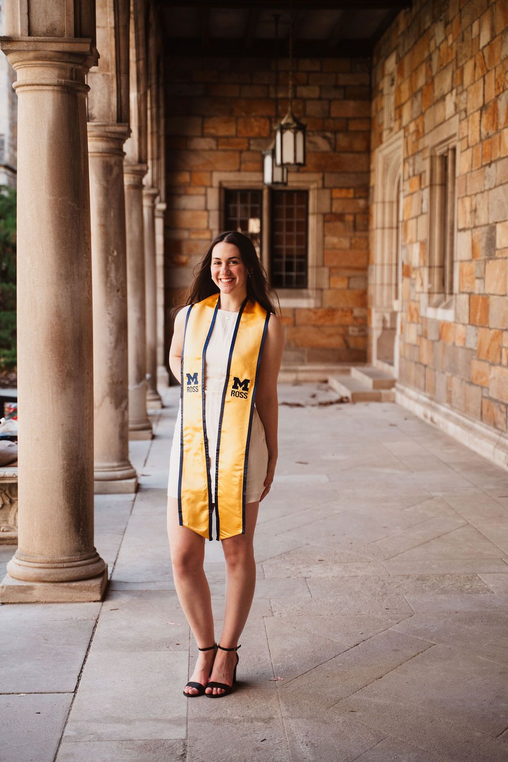 University of Michigan graduate smiling in gold graduation stole beside stone columns on campus.