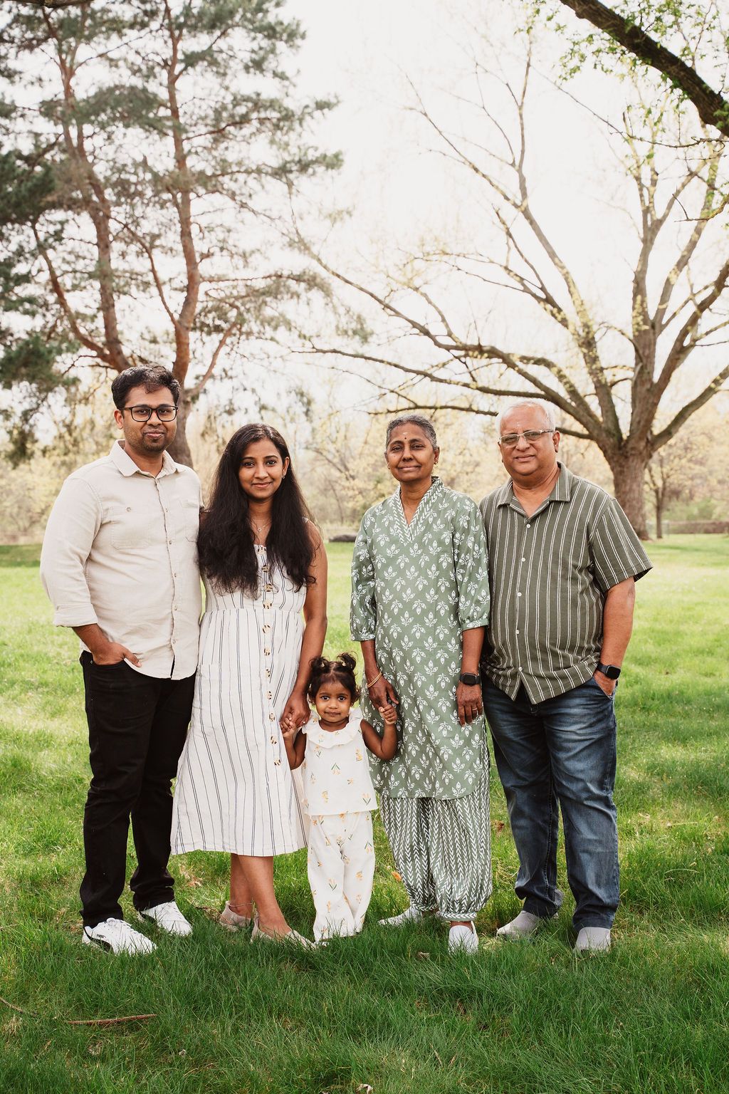 Multi-generational South Asian family of five posing outdoors in a lush green park with blooming trees.