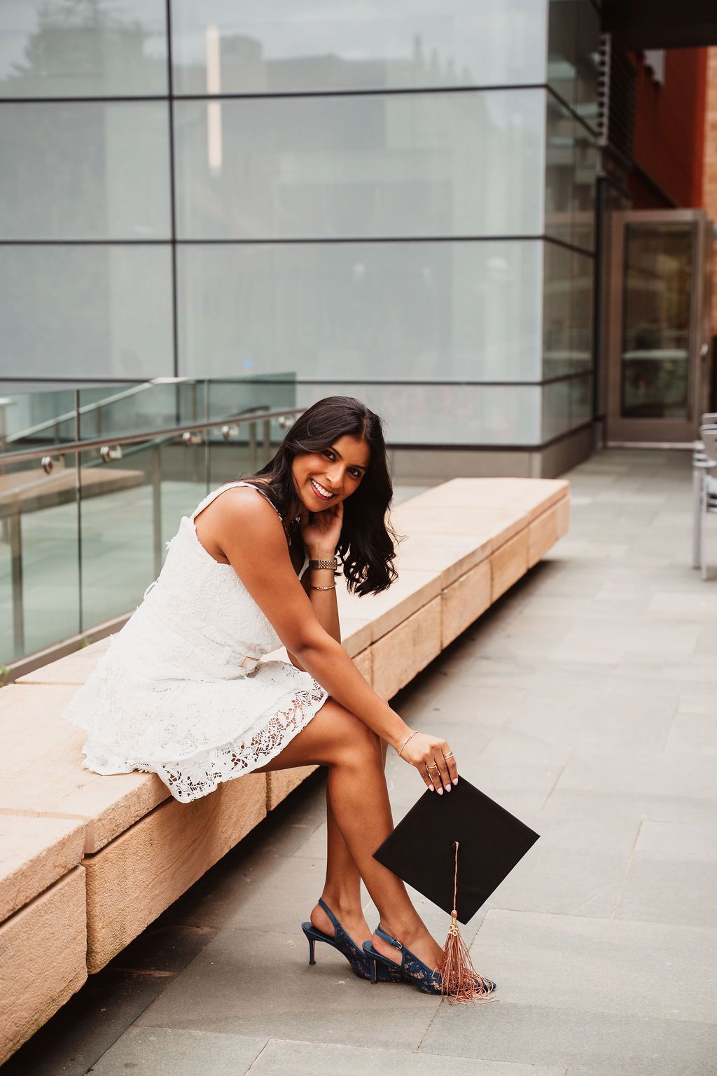 Smiling woman in white lace dress holding graduation cap, sitting on stone bench outside modern building.