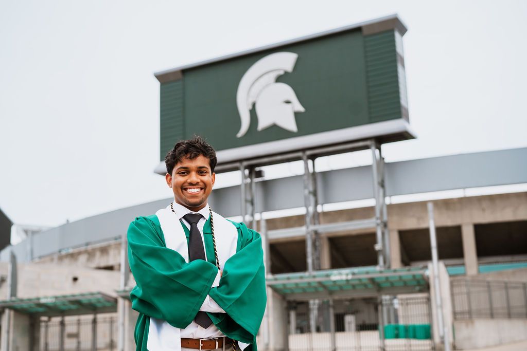 A graduate in green and white regalia poses outdoors near a building with a Spartan logo on the wall.
