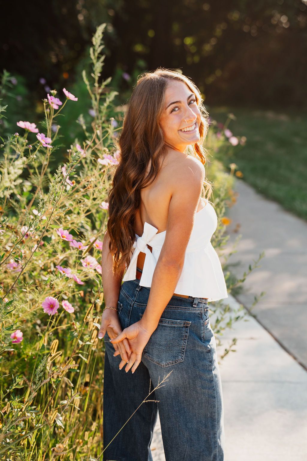 Smiling young woman with long wavy hair in white crop top and jeans posing by pink wildflowers on a sunny path.
