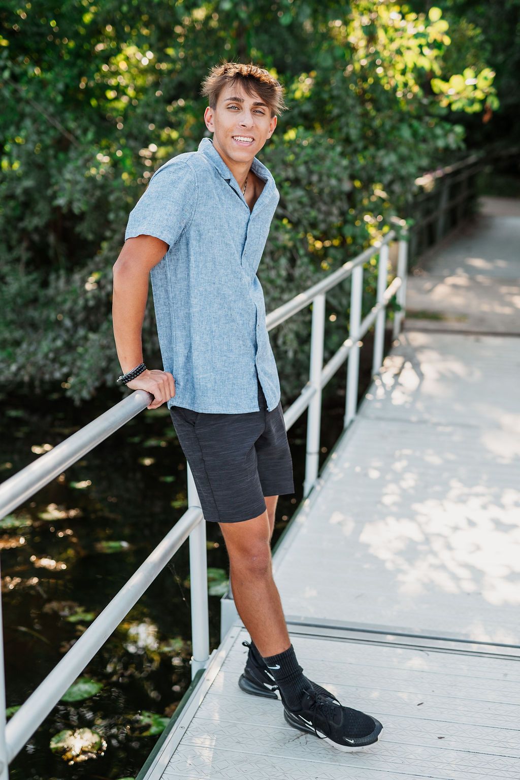 Young man in blue shirt and dark shorts smiling on a wooden bridge surrounded by lush green trees.