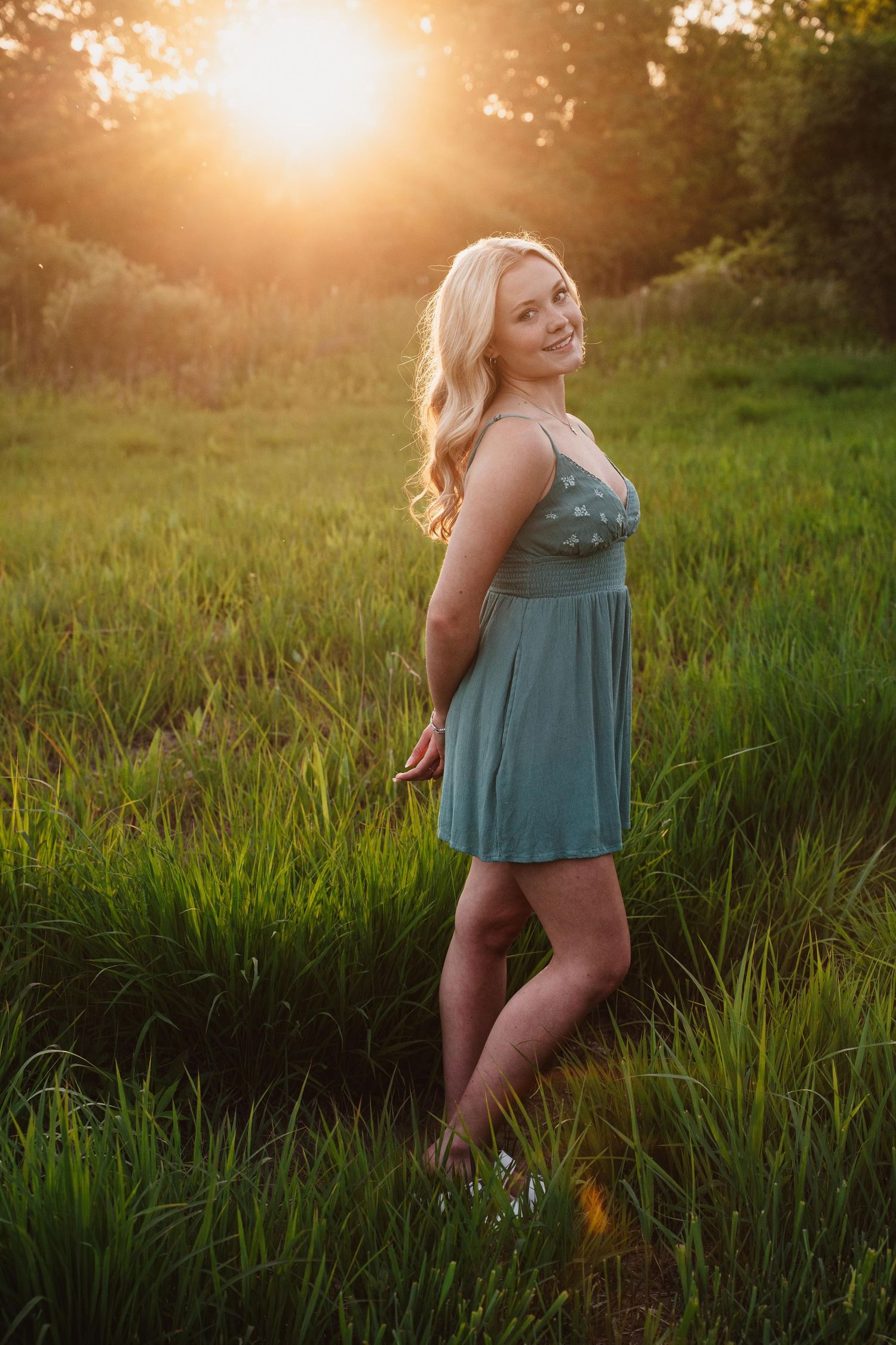 Blonde woman in teal dress standing in green field during golden sunset, smiling over shoulder.