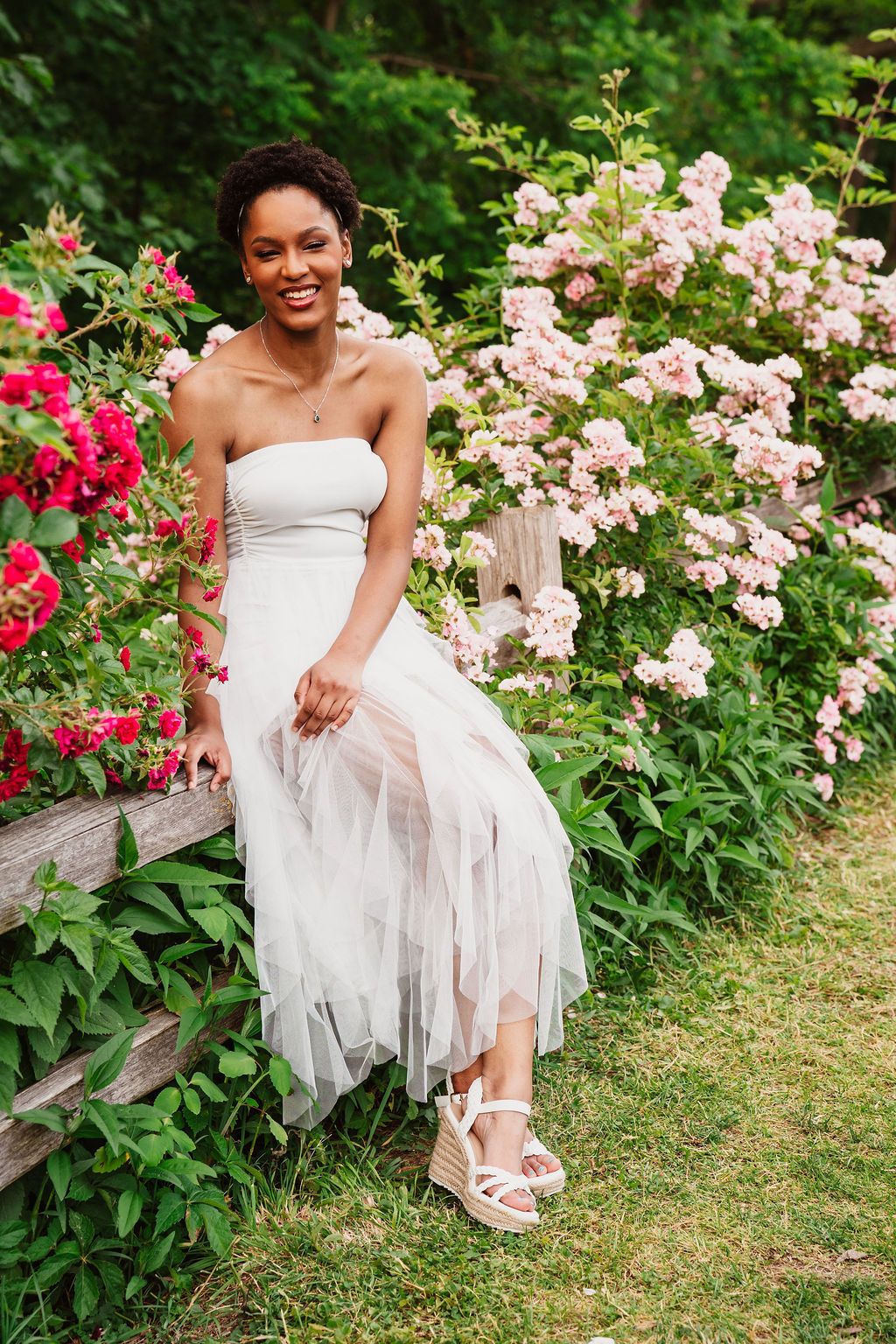 Smiling woman in white strapless tulle dress sits on wooden fence surrounded by blooming pink and red roses in garden.