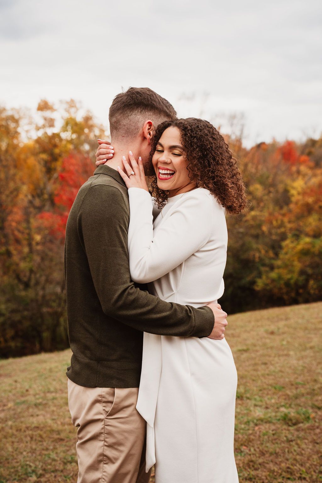 A joyful couple embraces in an autumn field, the woman in white laughing as colorful fall foliage fills the background.