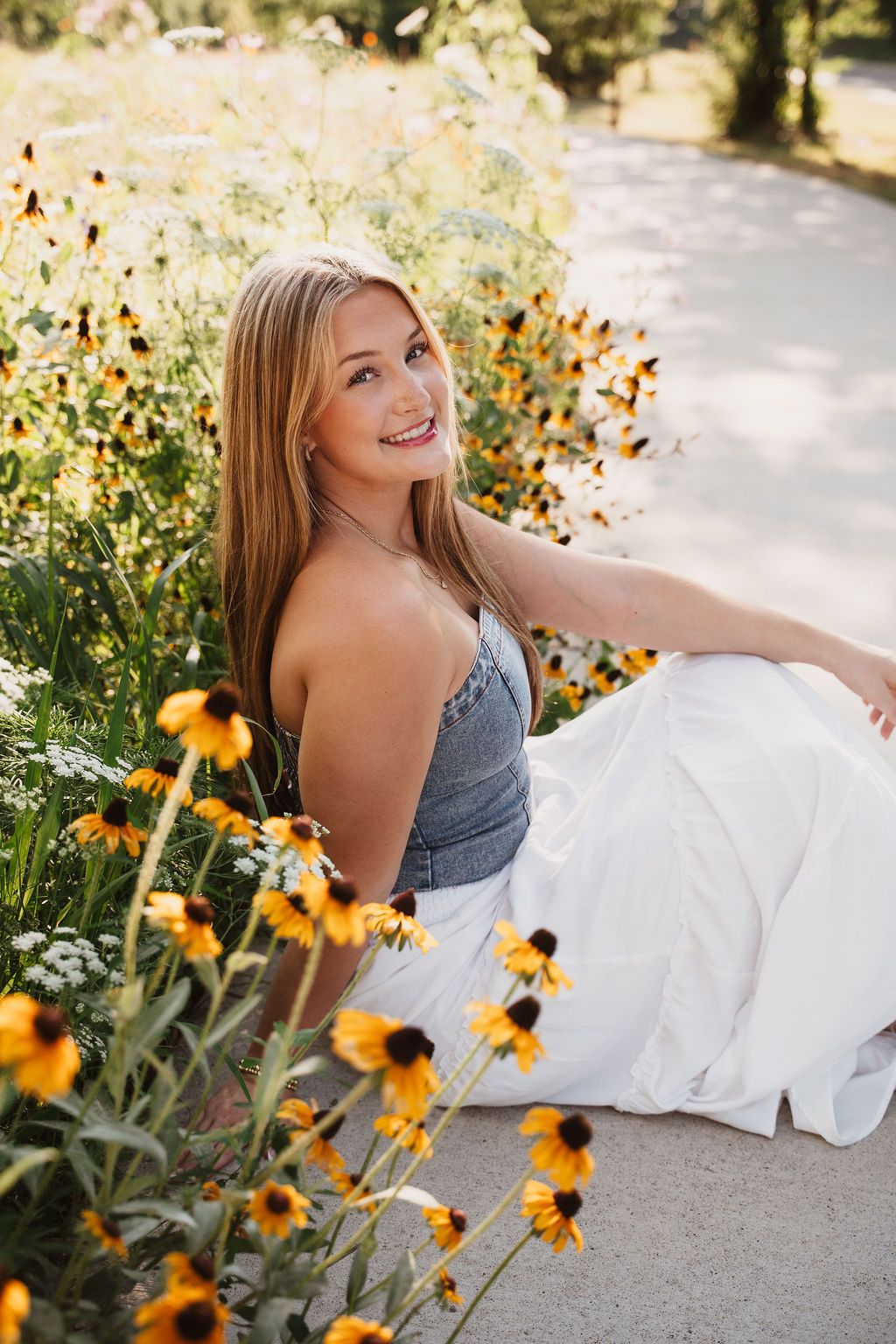 Smiling blonde woman in denim top and white skirt sitting among yellow black-eyed Susan wildflowers outdoors.