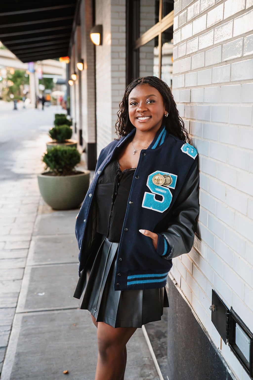 Student wearing a varsity letterman jacket poses confidently against a white brick wall.