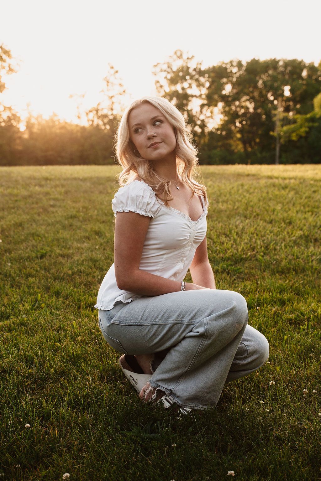 Young blonde woman in white top and jeans sits cross-legged on green grass during golden hour sunset.