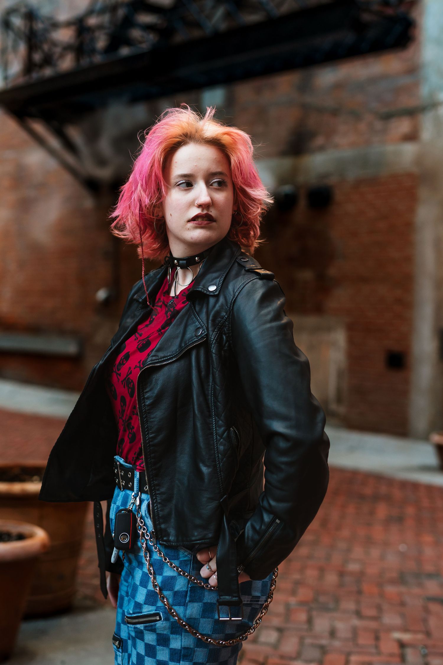 Young woman with pink hair wearing a black leather jacket and checkered pants poses in an urban brick alleyway.