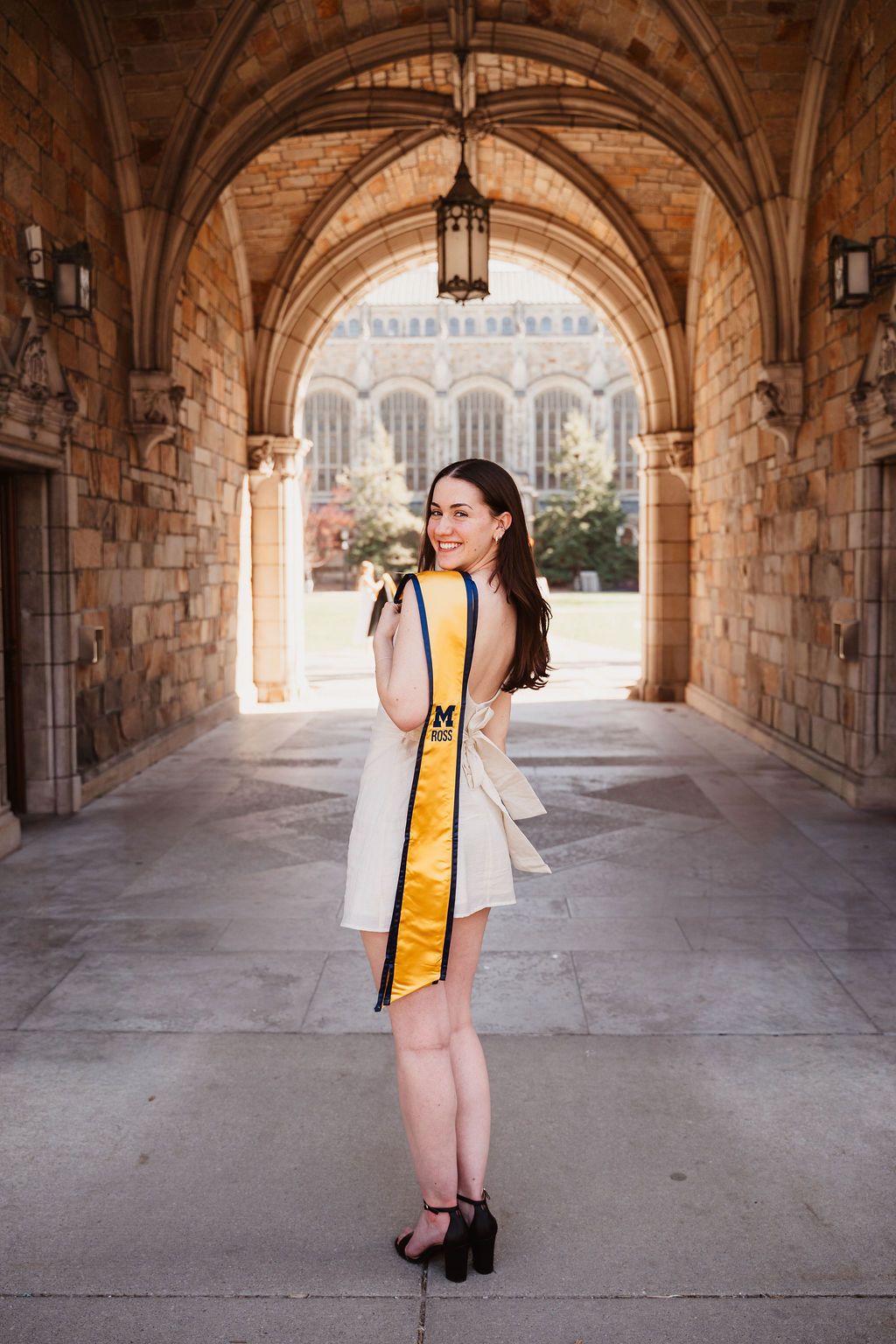 Smiling graduate in white dress with gold University of Michigan sash poses under stone archway on campus.
