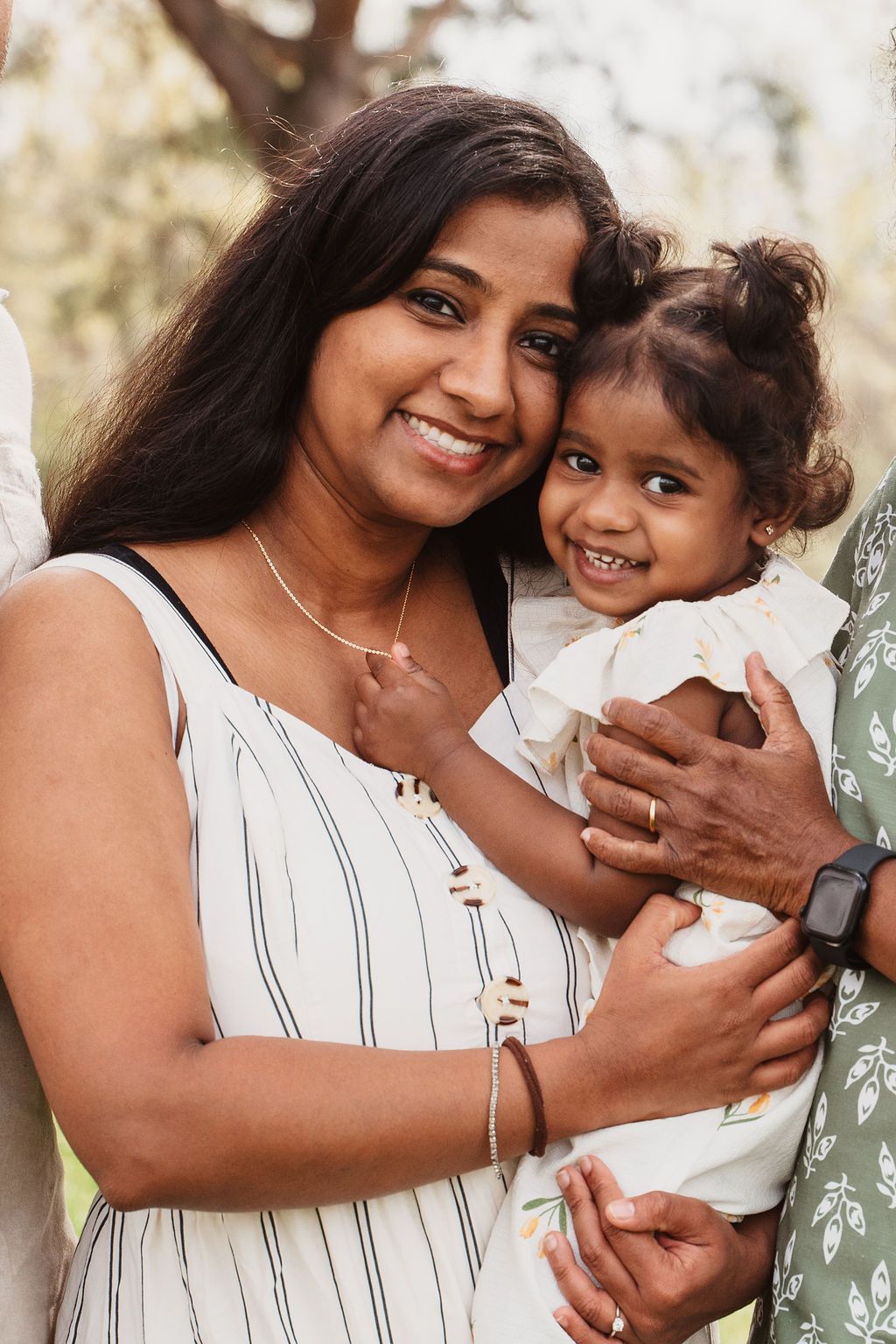 A smiling woman in a striped dress holds a happy toddler girl outdoors in a sunlit park setting.