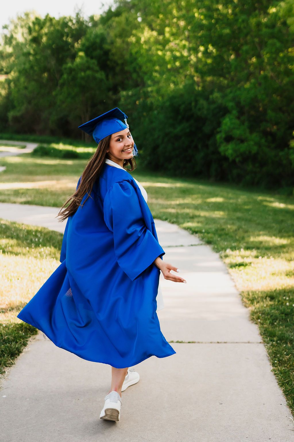Graduate in bright blue cap and gown twirls joyfully on a sunny pathway during graduation photo session.