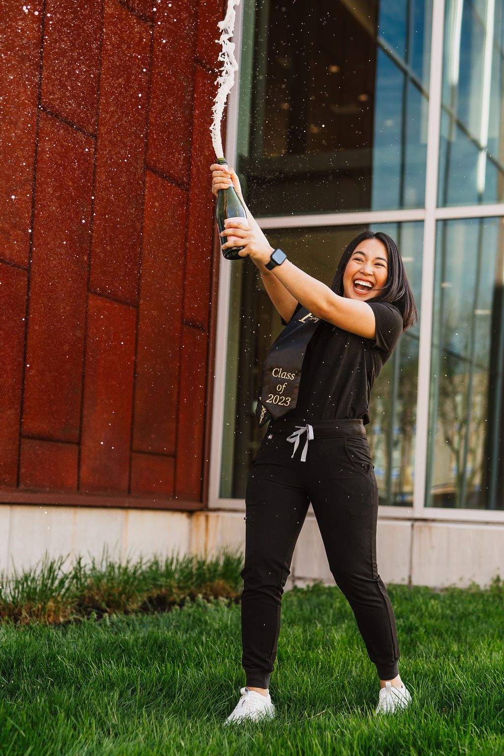 Woman in black athleisure outfit poses playfully on green grass outside modern building with red walls.