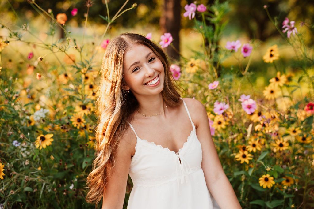 A person in a white sundress smiles while standing in a garden of yellow and pink wildflowers on a sunny day.