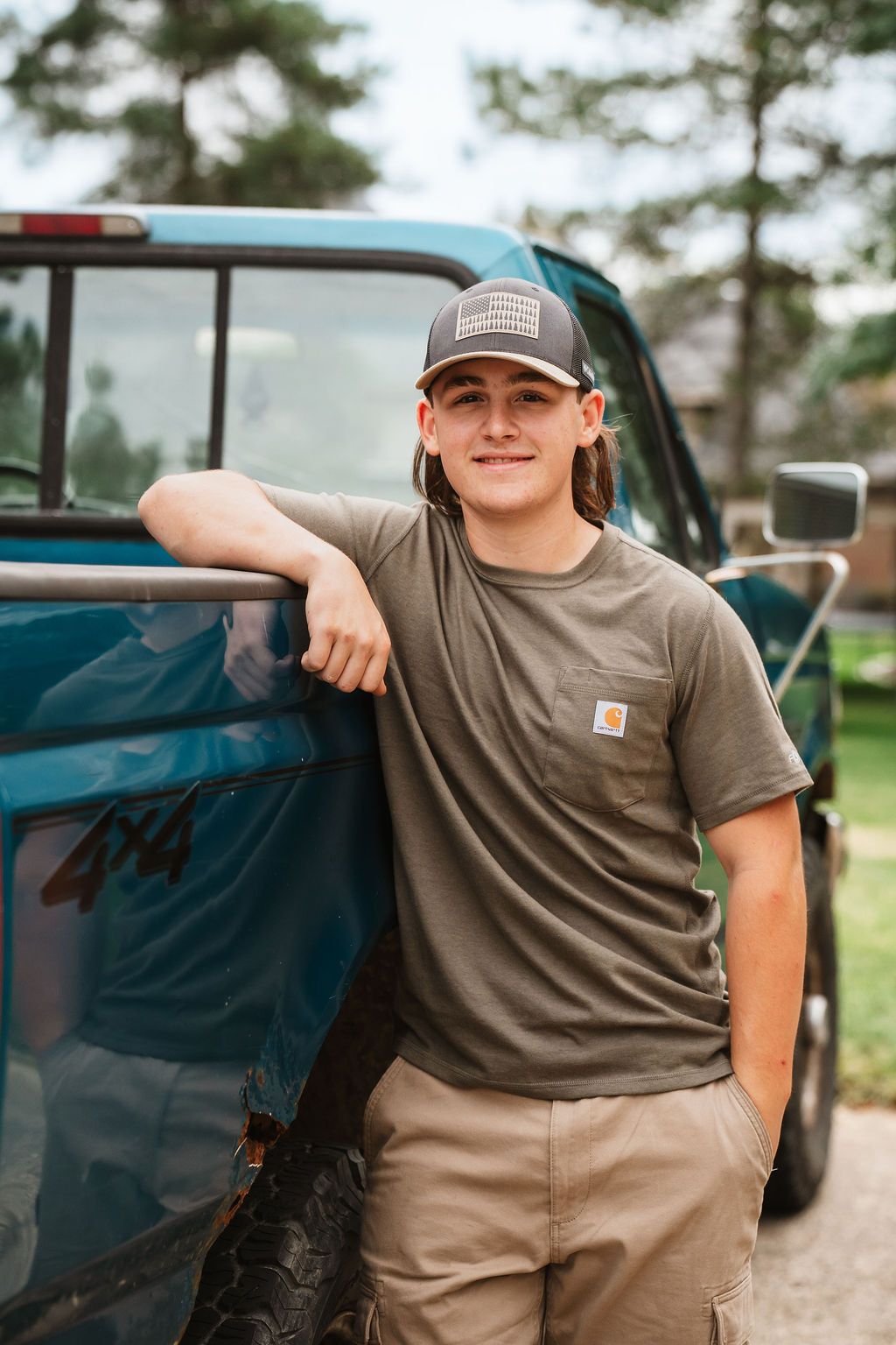 Person wearing a Carhartt t-shirt and baseball cap leans against a blue pickup truck in an outdoor setting.
