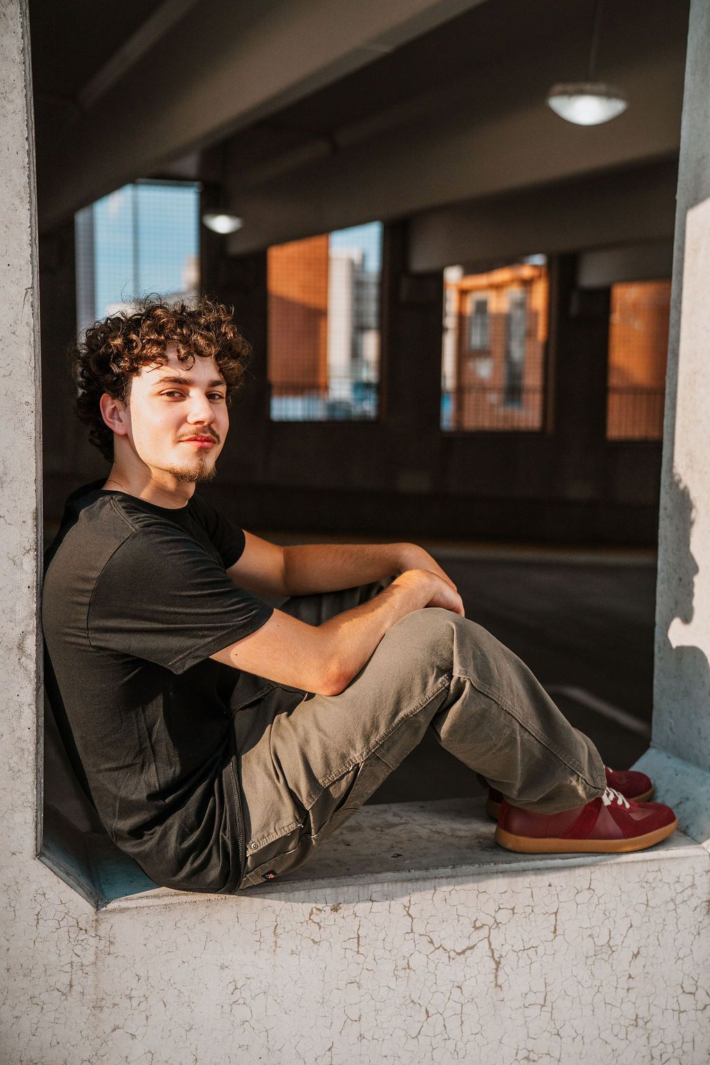 Young man with curly hair sits relaxed in urban parking garage, wearing black tee, khaki pants, and red sneakers.