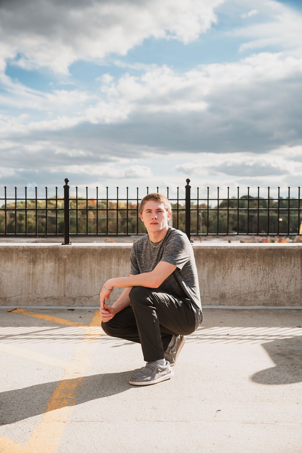 Young man crouching on rooftop parking lot, wearing gray t-shirt and black pants, with cloudy sky backdrop.