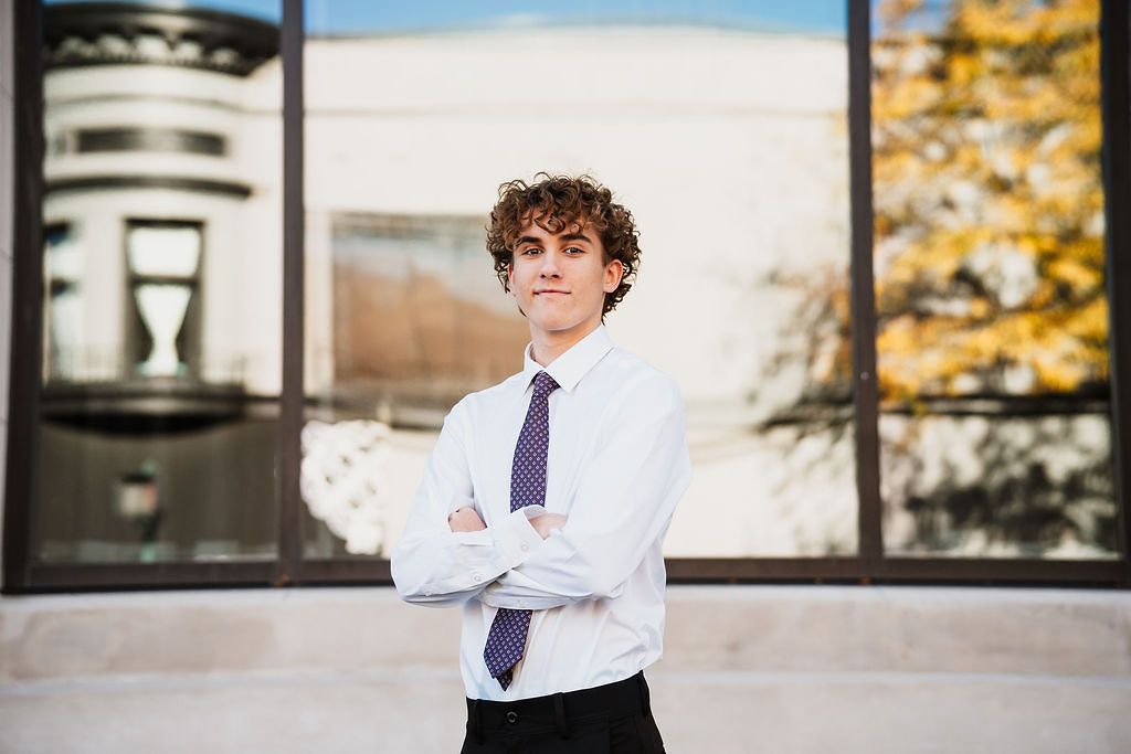 Young professional man in white shirt and purple tie stands confidently with arms crossed outdoors.