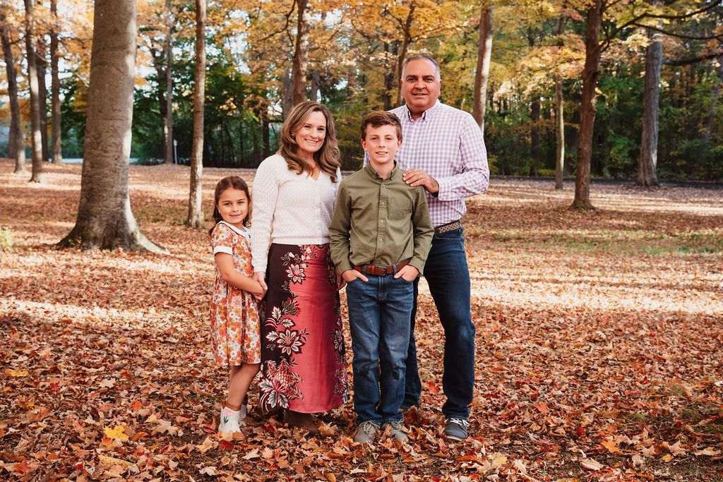 Family of four smiling together in autumn park with colorful fall foliage and leaves on the ground.