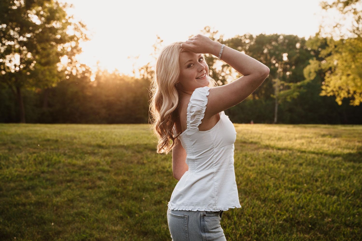 Young blonde woman in white top and jeans poses in sunlit green field during golden hour sunset.