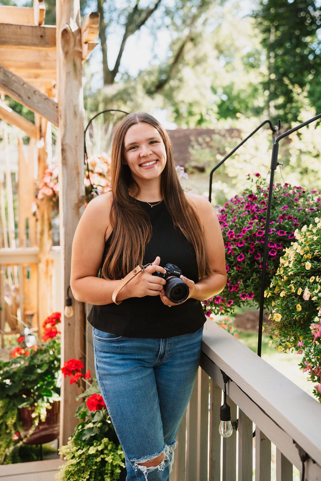 Photographer in black top and jeans poses with camera on wooden porch with flowers.