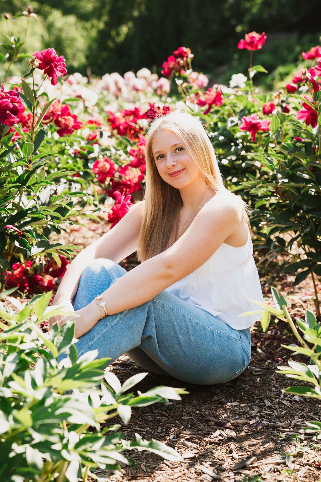 Person in white top and jeans sitting among vibrant red roses in a garden.
