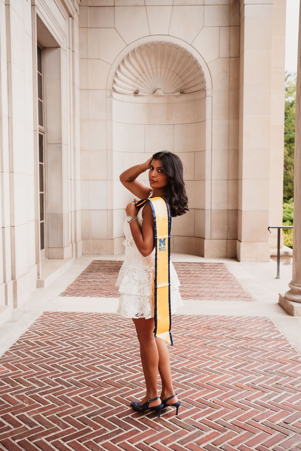 Young woman in white dress and gold graduation stole poses on brick walkway at university building.