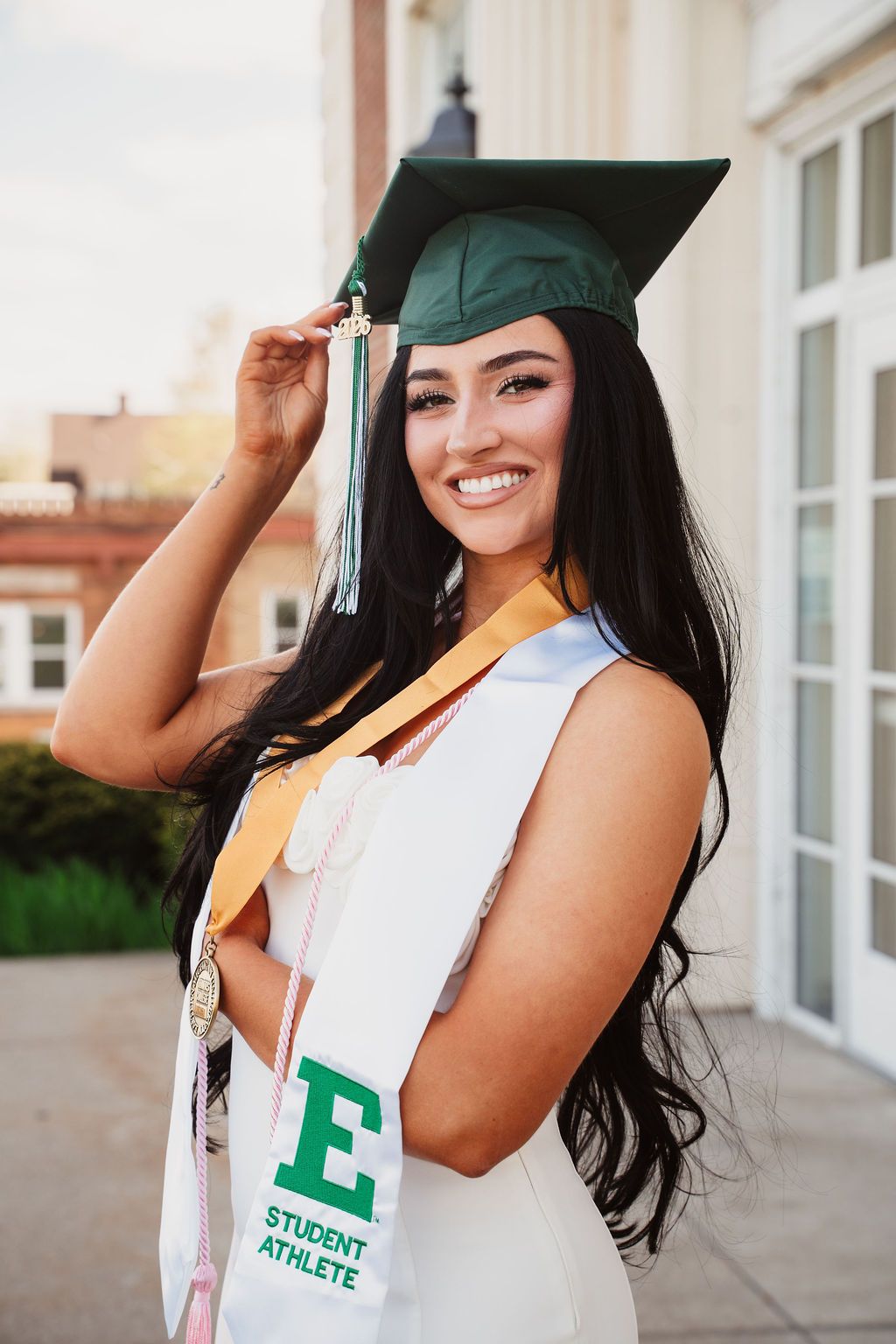 Smiling female graduate in green cap and gown with Eastern Michigan University Student Athlete sash.