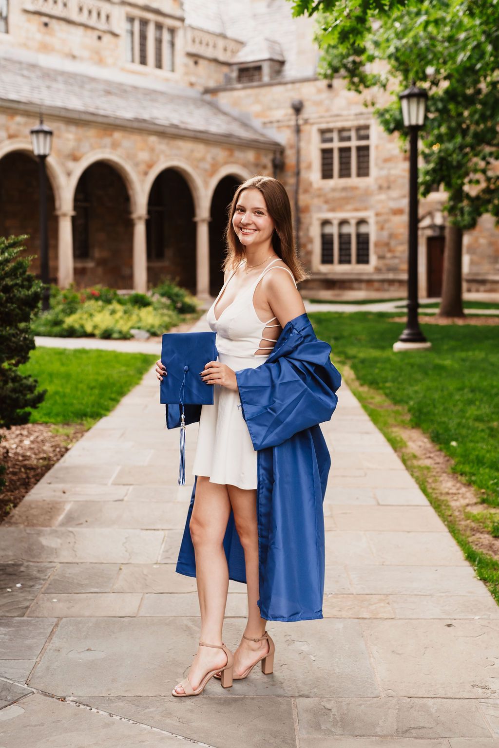 Graduate in white dress and blue graduation gown stands on stone path at historic university campus with arches.