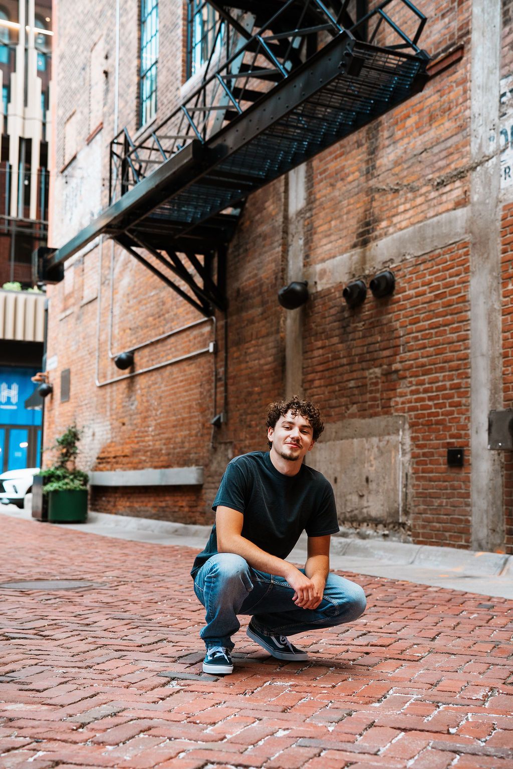 Person sitting on red brick cobblestone alley with rustic brick wall and fire escapes in urban setting.
